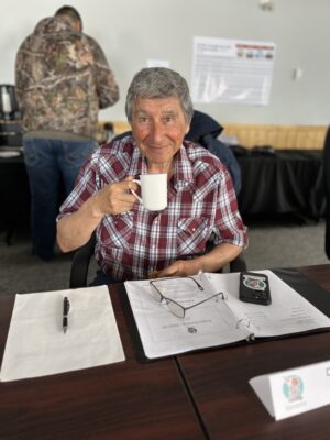 A man in a plaid shirt sits at a table smiling, holding a cup of coffee with notebooks open in front of him.
