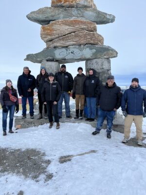 Nine people stand on snowy ground in front of an inukshuk.