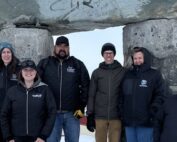 Nine people stand on snowy ground in front of an inukshuk.