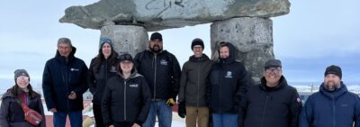 Nine people stand on snowy ground in front of an inukshuk.