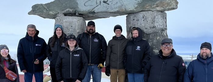 Nine people stand on snowy ground in front of an inukshuk.